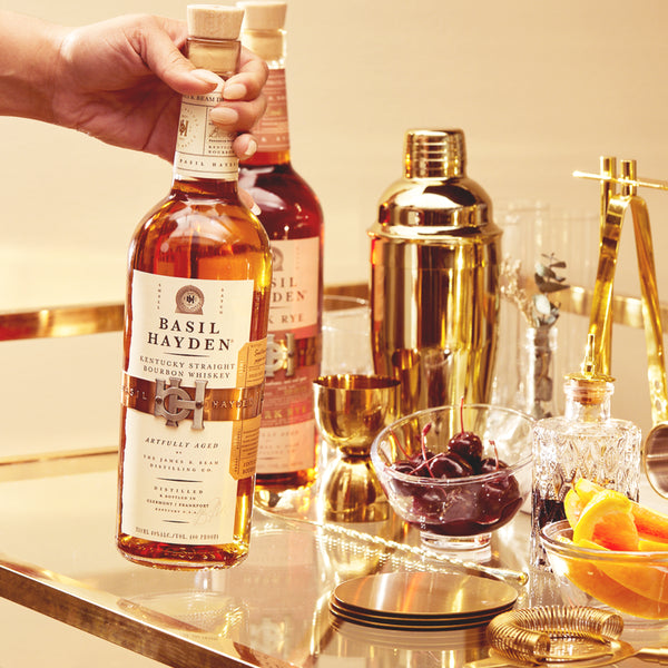 Load image into Gallery viewer, Bottle of Basil Hayden whiskey being held over a bar setup with glasses and ingredients.