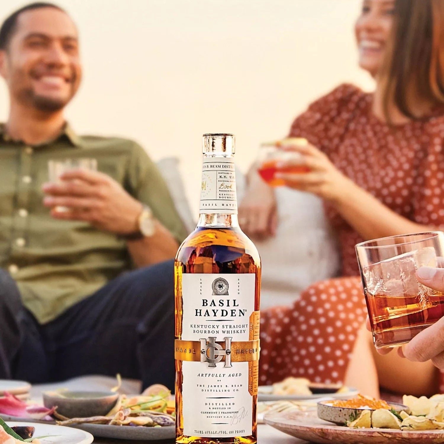 Load image into Gallery viewer, Bottle of Basil Hayden's whiskey on a table with people in the background.