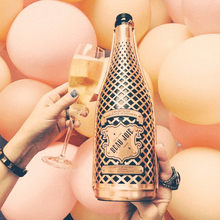 Load image into Gallery viewer, Person holding a bottle of Beau Joie Champagne bottle and glass against a pink balloon background.