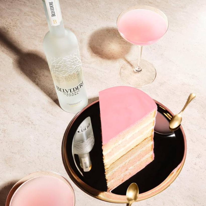 Layered cake with pink and white frosting on a black plate, accompanied by a bottle of Belvedere vodka and a pink cocktail glass.