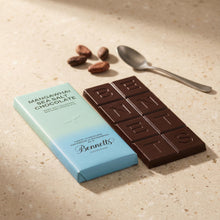 Load image into Gallery viewer, Bennett's chocolate bar and packaging on a textured surface with cocoa beans and a spoon.