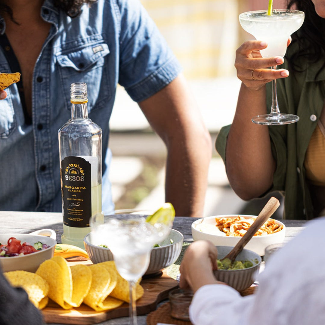 Load image into Gallery viewer, People enjoying a meal with a bottle of Besos Margarita Clasica ready to serve cocktail on a table.