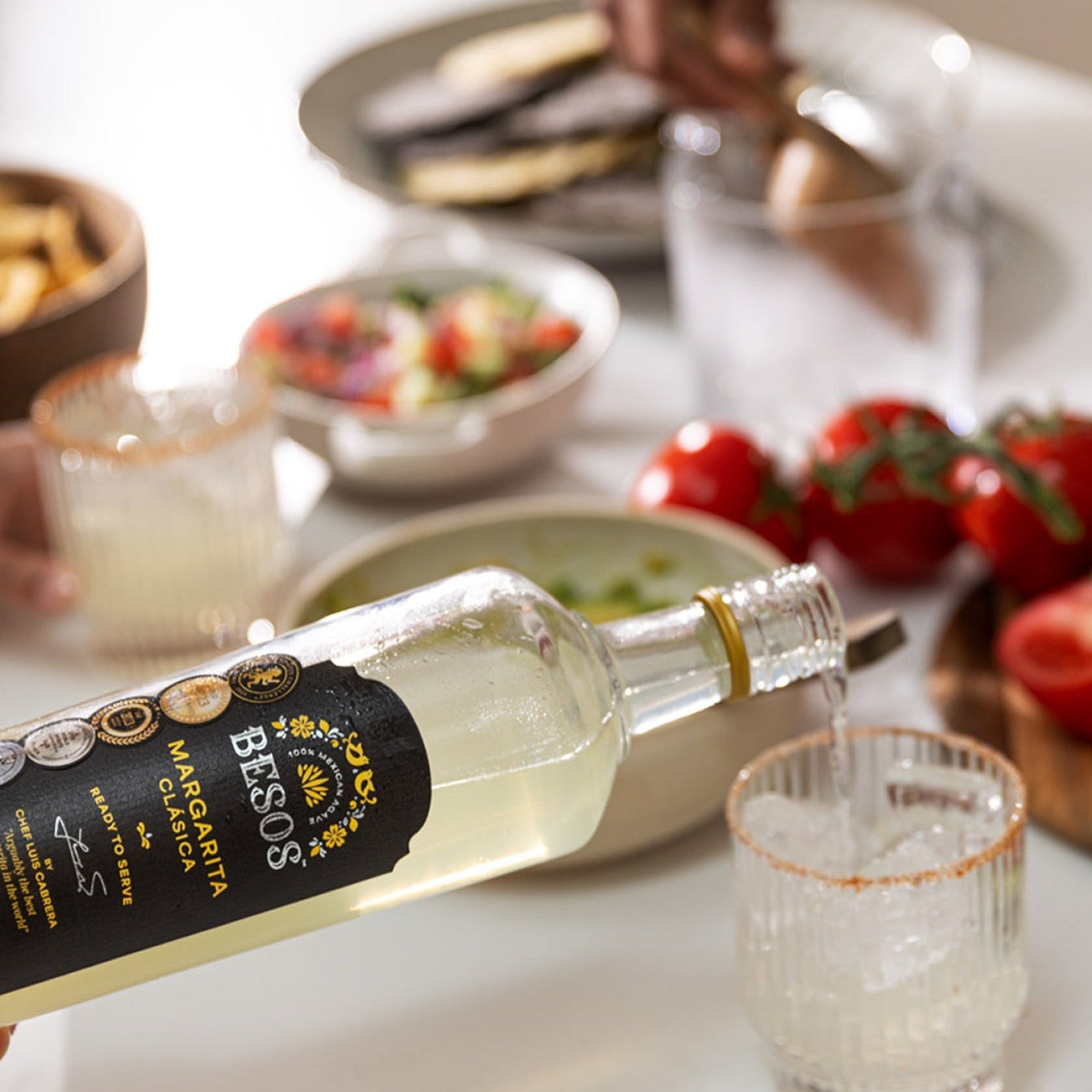 Load image into Gallery viewer, Bottle of Besos Margarita Clasica ready to serve cocktail with a glass on a table with food and fruit.
