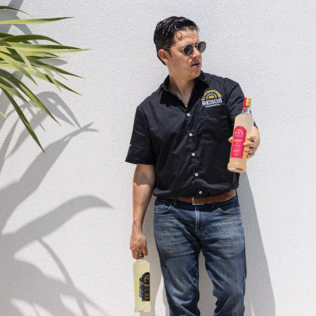 Load image into Gallery viewer, Luis holding two bottles of Besos Margarita ready to serve cocktails against a white wall with a plant to the left.