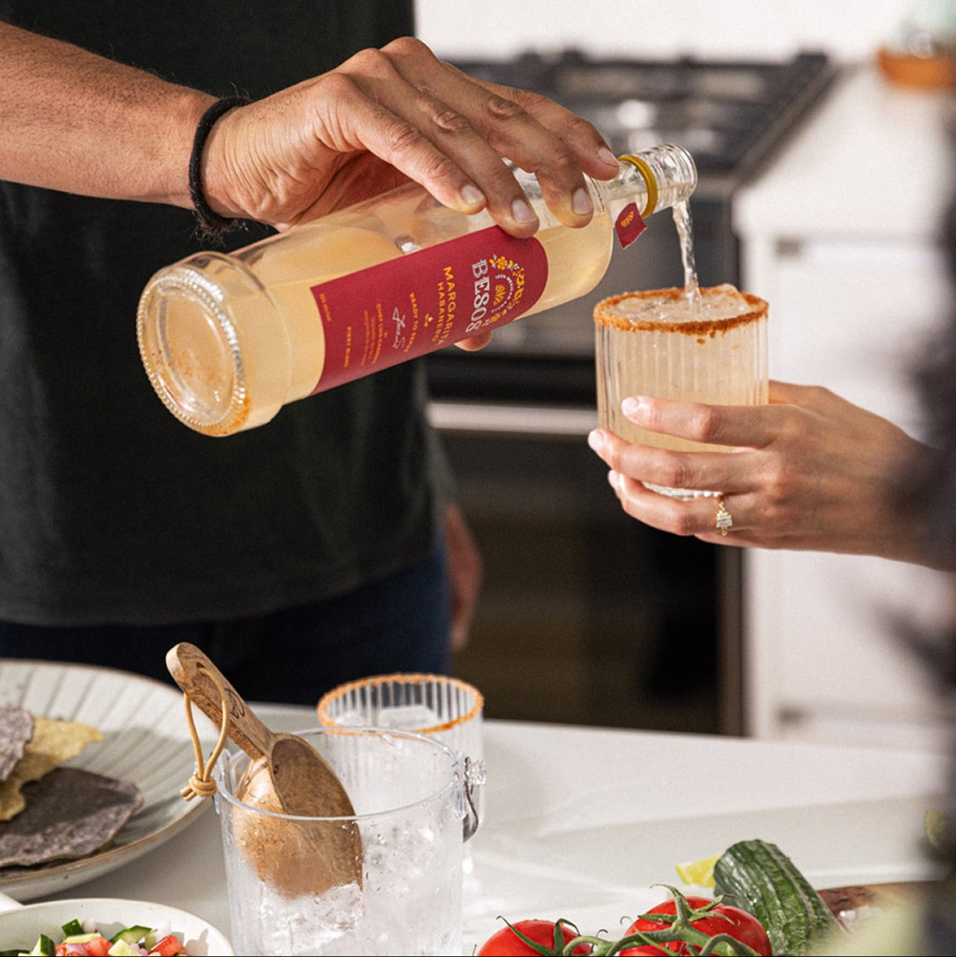 Load image into Gallery viewer, Person pouring a Besos Margarita Habanero ready to serve spicy cocktail  from a bottle into a glass with food on a table in the background