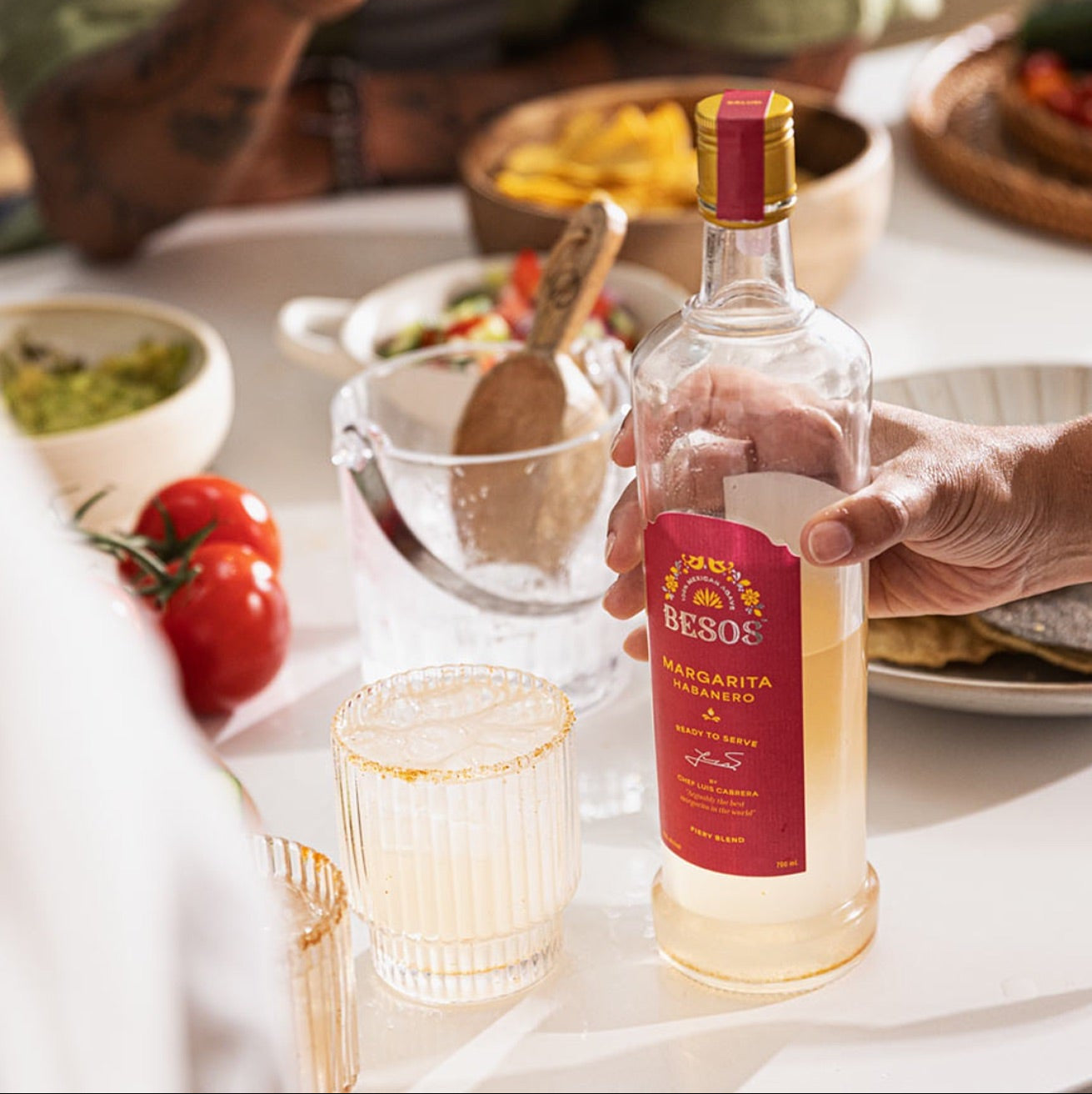 Load image into Gallery viewer, Person holding a bottle of Besos Margarita Habanero ready to serve spicy cocktail  with a table setting in the background