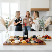 Load image into Gallery viewer, Three women in a kitchen enjoying a bottle of Bilancia La Collina Syrah wine and a charcuterie board with fruits and cheeses.