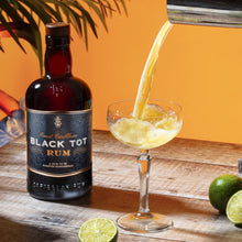 Load image into Gallery viewer, Black Tot Rum being poured into a glass with limes and books on a wooden table.