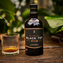 Load image into Gallery viewer, Black Tot Rum bottle with a glass of rum on a wooden surface with greenery in the background