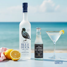 Load image into Gallery viewer, Bottle of Blue Duck vodka, bottle of East Imperial soda water, and cocktail with lemon slice on a table with ocean view.