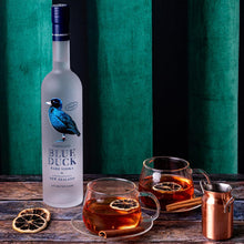 Load image into Gallery viewer, Bottle of Blue Duck vodka with two glasses of tea on a wooden surface against a green curtain.