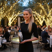 Load image into Gallery viewer, Woman holding a bottle of Bogle Phantom Proprietary Red Blend wine in an outdoor setting with festive lights and people in the background