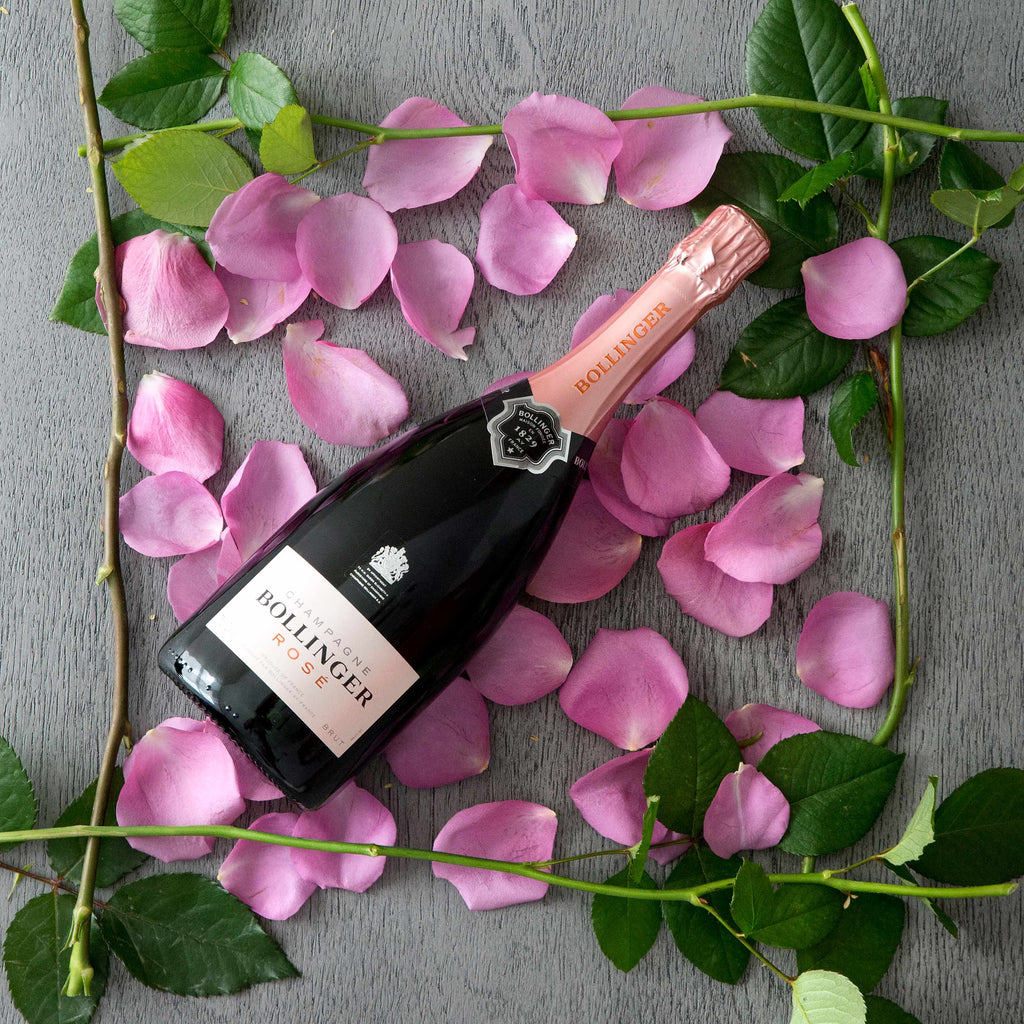 Bollinger rosé champagne bottle surrounded by pink petals and green leaves on a gray surface