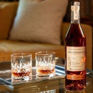 A bottle of Bombergers bourbon on a glass table with two glasses in a cozy living room setting.