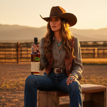 Load image into Gallery viewer, Cowgirl holding a bottle of Bookers-Bourbon-The-Lumberyard-Batch-Whiskey  with a scenic background