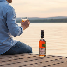 Load image into Gallery viewer, Person sitting by a lake holding a glass of whiskey next to a bottle of Bookers-Bourbon-The-Lumberyard-Batch-Whiskey on a wooden dock.