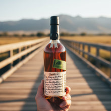 Load image into Gallery viewer, Bottle of Bookers-Bourbon-The-Lumberyard-Batch-Whiskey held in front of a scenic wooden bridge with mountains in the background