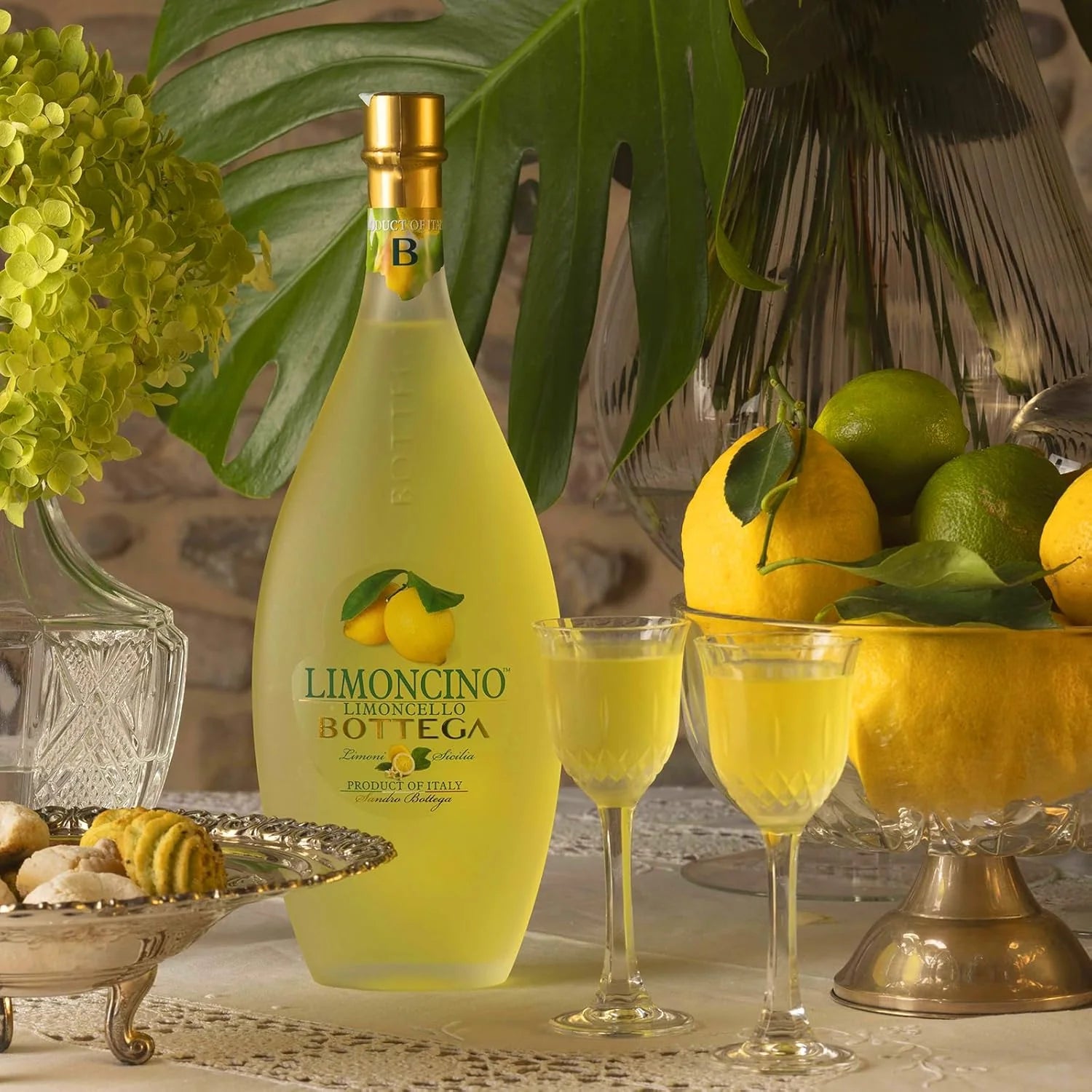 Load image into Gallery viewer, Bottle of Limoncino Bottega with two glasses filled with the product, surrounded by lemons and limes.