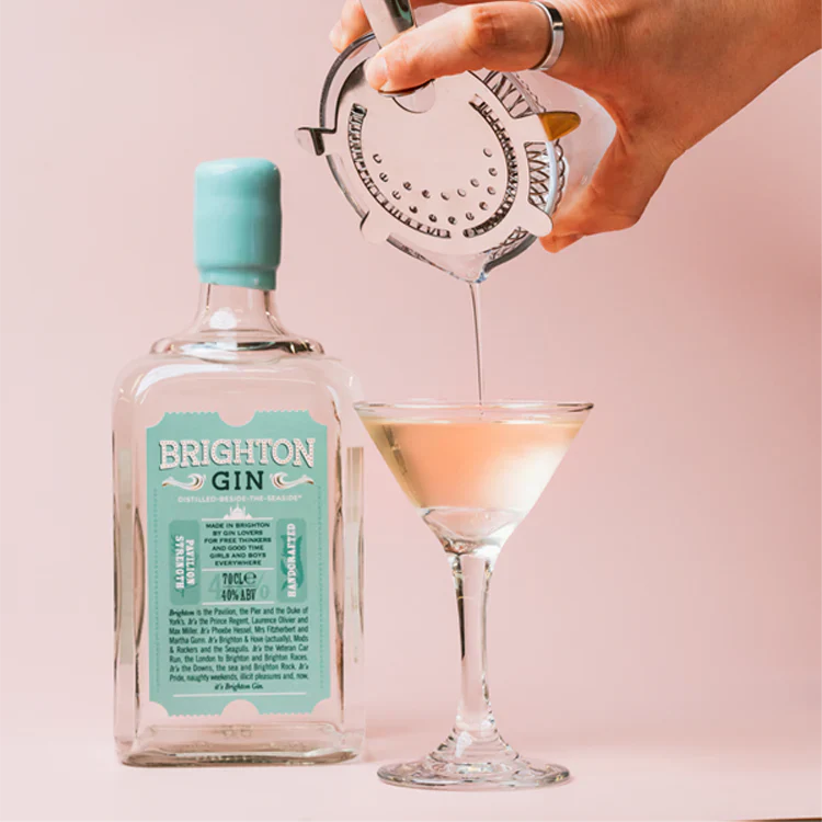 Load image into Gallery viewer, Person pouring a drink from a bottle labeled 'Brighton Gin' into a cocktail glass against a pink background.