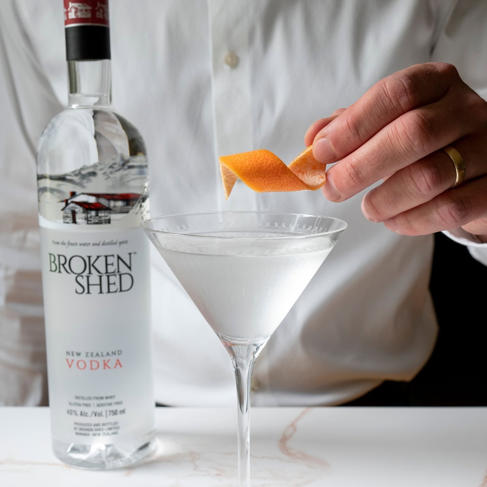Load image into Gallery viewer, A bar man in a white shirt garnishes a martini glass with an orange peel, with a bottle of Broken Shed New Zealand Vodka standing on a white marble surface beside it.
