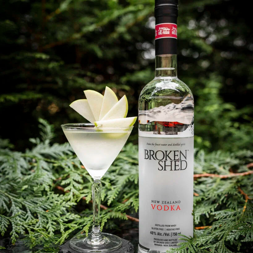 Load image into Gallery viewer, A bottle of Broken Shed New Zealand Vodka stands next to a martini glass filled with white cocktail liquor and garnished with a sliced pear, set against a dark green forest background with pine branches.