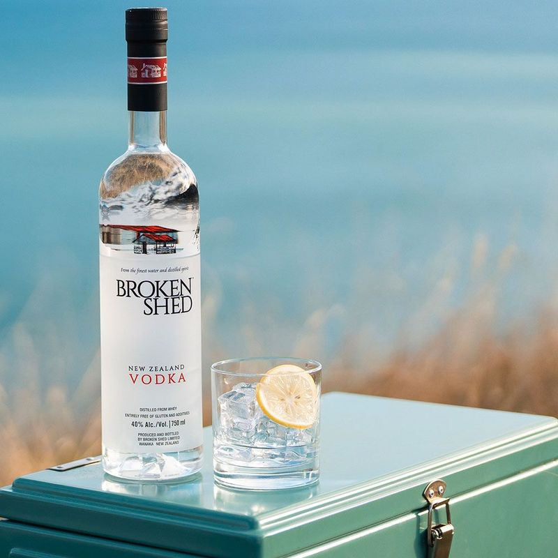 Load image into Gallery viewer, A bottle of Broken Shed New Zealand Vodka stands on a teal cooler beside a glass of water with a lemon slice, with a blurred beach or water background.