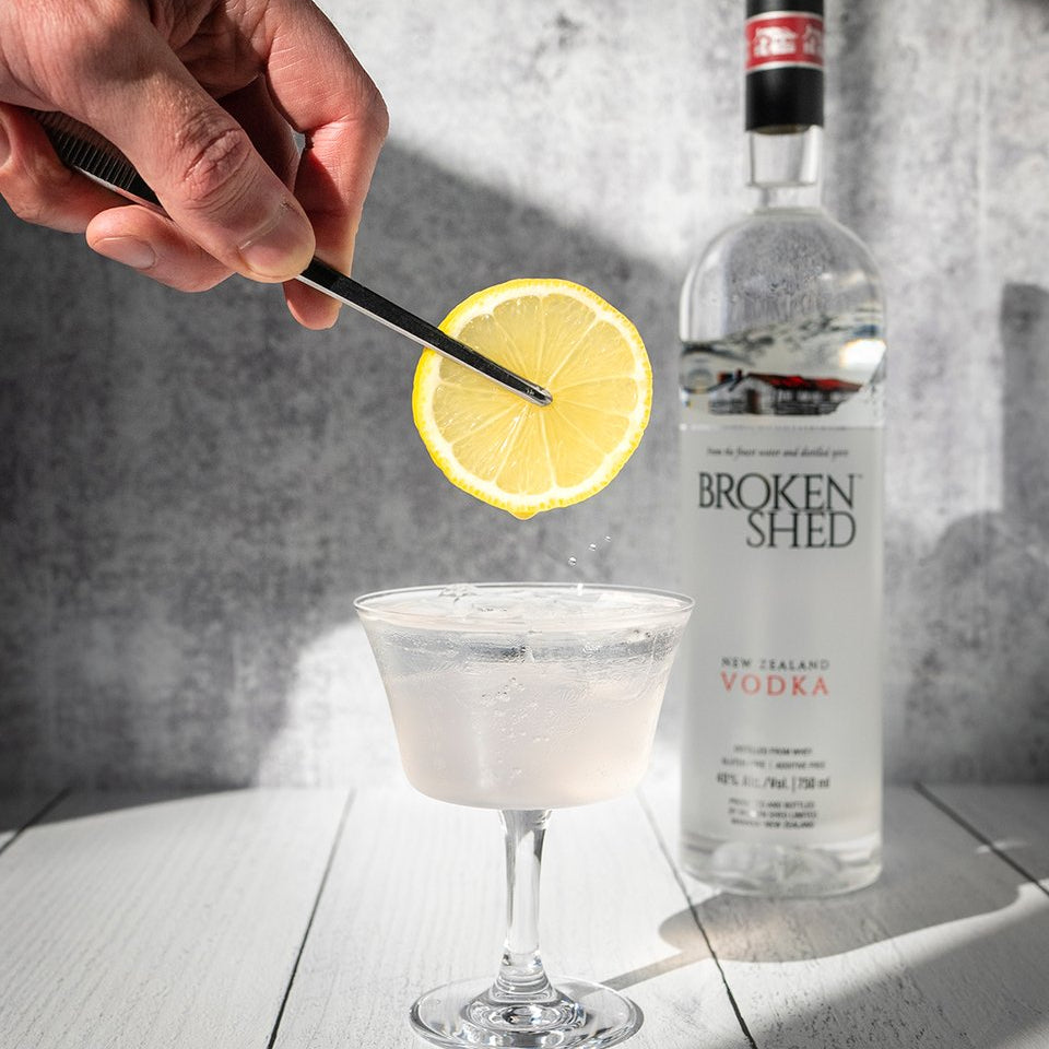 Load image into Gallery viewer, A hand holds a lemon slice above a filled coupe glass, with a bottle of Broken Shed New Zealand Vodka standing behind on a white wood surface against a grey textured wall.