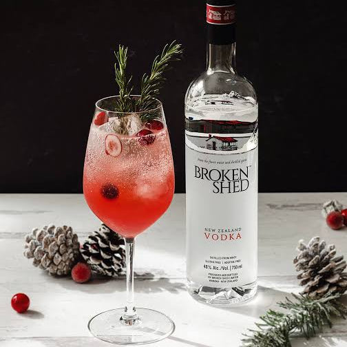 Load image into Gallery viewer, A bottle of Broken Shed New Zealand Vodka stands next to a pink cocktail in a stemmed glass, decorated with a rosemary sprig and cranberry slices, set on a white marble surface with pinecones and cranberries against a dark background.