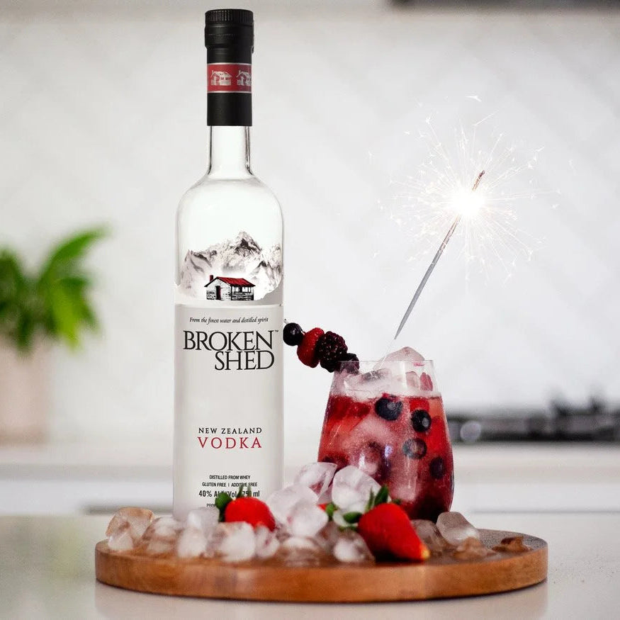 Load image into Gallery viewer, A bottle of Broken Shed New Zealand Vodka stands on a wooden tray beside a glass of red cocktail with ice, berries, and a sparkler in a bright kitchen setting.