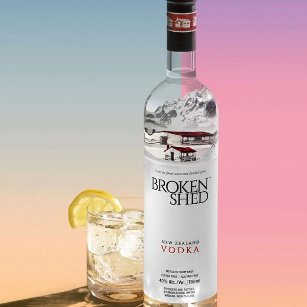 Load image into Gallery viewer, A bottle of Broken Shed New Zealand Vodka stands next to a glass of mixed drink with ice and a lemon slice, set against a pastel gradient background.