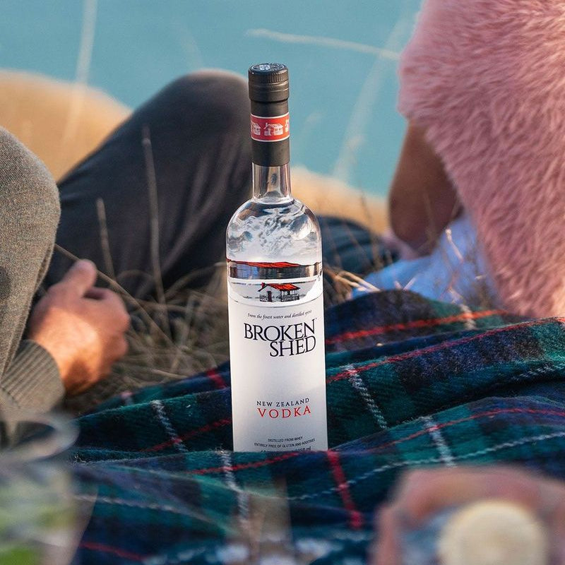 Load image into Gallery viewer, A bottle of Broken Shed New Zealand Vodka stands on a plaid blanket outdoors, with people seated around it in a casual, warm setting.