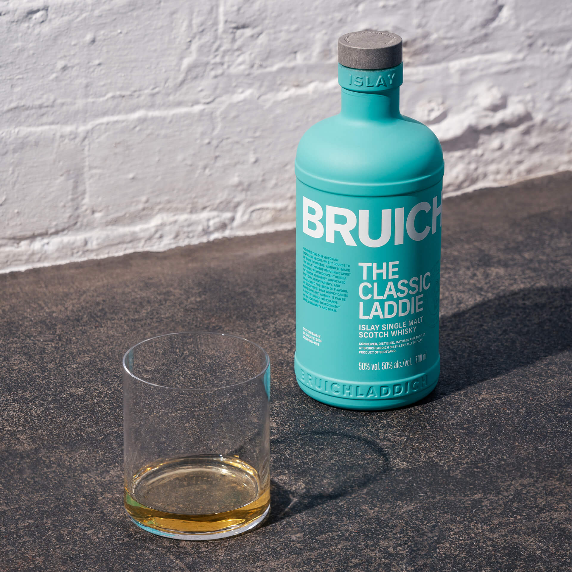 Load image into Gallery viewer, Bruichladdich whisky bottle with a glass on a stone surface against a white brick wall.