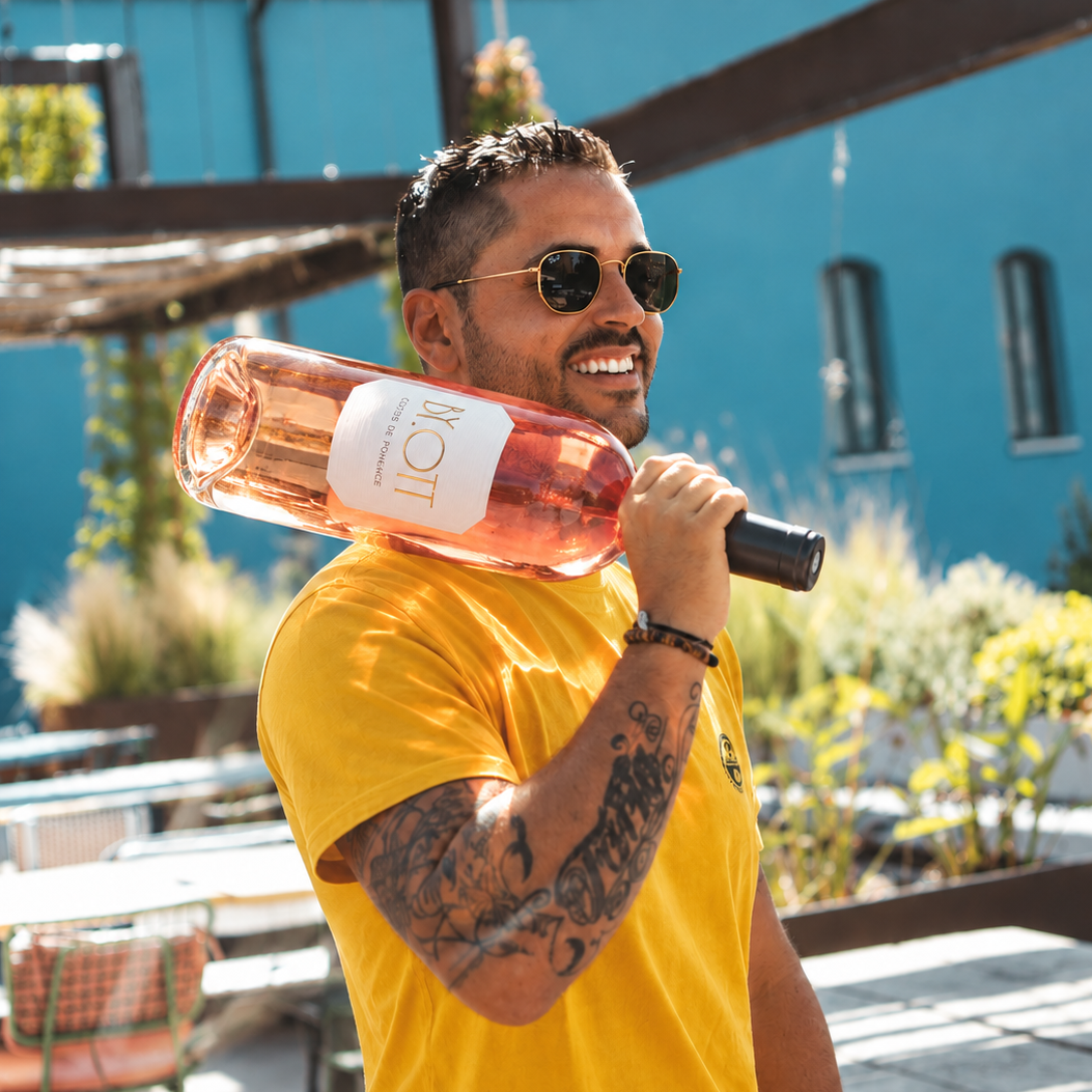 Load image into Gallery viewer, A man in a yellow t-shirt and sunglasses holds a large bottle of Jeroboam 3000ml BY.OTT Rosé wine over his shoulder in an outdoor patio setting.