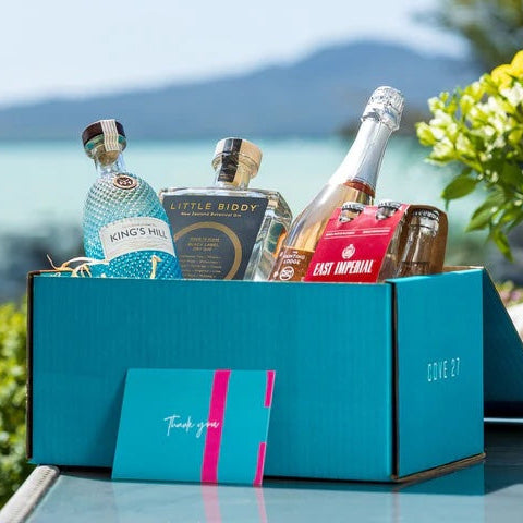 Load image into Gallery viewer, A teal coloured gift box with a couple of bottles of gin, a champagne and some tonics in it with a background of the sea and a mountain