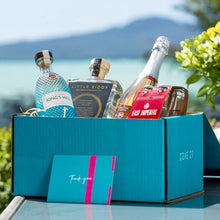 Load image into Gallery viewer, A teal coloured gift box with a couple of bottles of gin, a champagne and some tonics in it with a background of the sea and a mountain