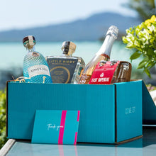 Load image into Gallery viewer, A teal coloured gift box with a couple of bottles of gin, a champagne and some tonics in it with a background of the sea and a mountain