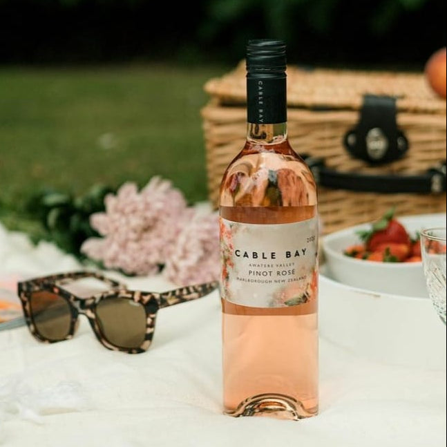Load image into Gallery viewer, Bottle of Cable Bay Rosé wine on a picnic blanket with sunglasses and flowers in the background