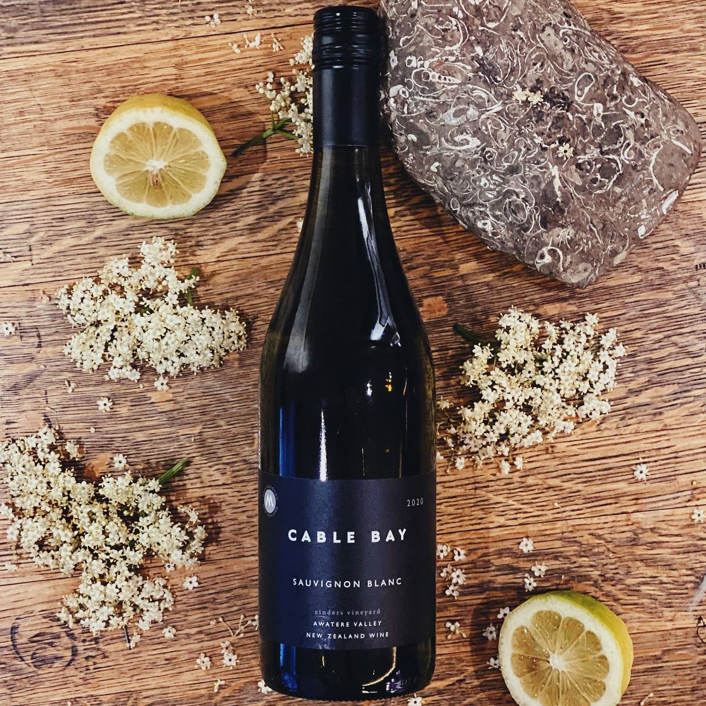 Load image into Gallery viewer, A bottle of Cable Bay Cinders Vineyard Sauvignon Blanc on a wooden surface with some blowers and lemons around it
