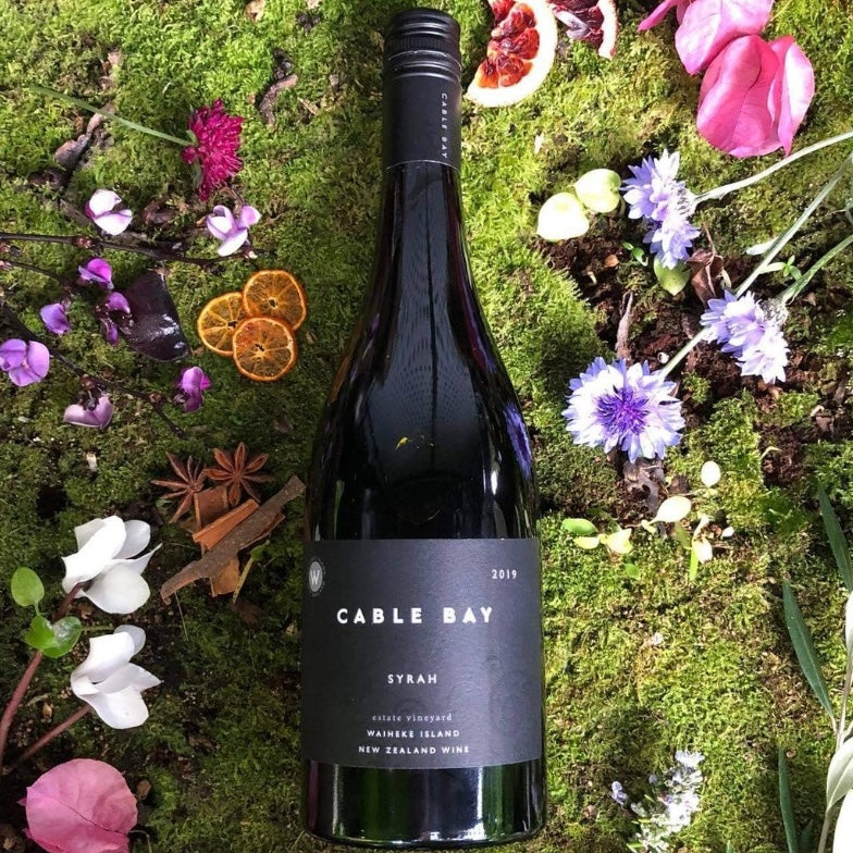 Load image into Gallery viewer, a bottle of Cable Bay Waiheke Island Syrah on the grass with citrus and flower petals surrounding it.