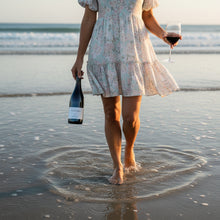Load image into Gallery viewer, Person in a floral dress walking on the beach with a bottle of Cable Bay Reserve Syrah and glass.