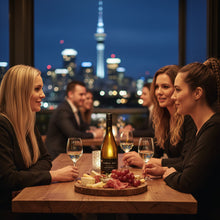 Load image into Gallery viewer, Three women in their 30s in a restaurant enjoying a bottle of Cable Bay Waiheke Island Chardonnay wine with the Auckland Sky Tower in the background.