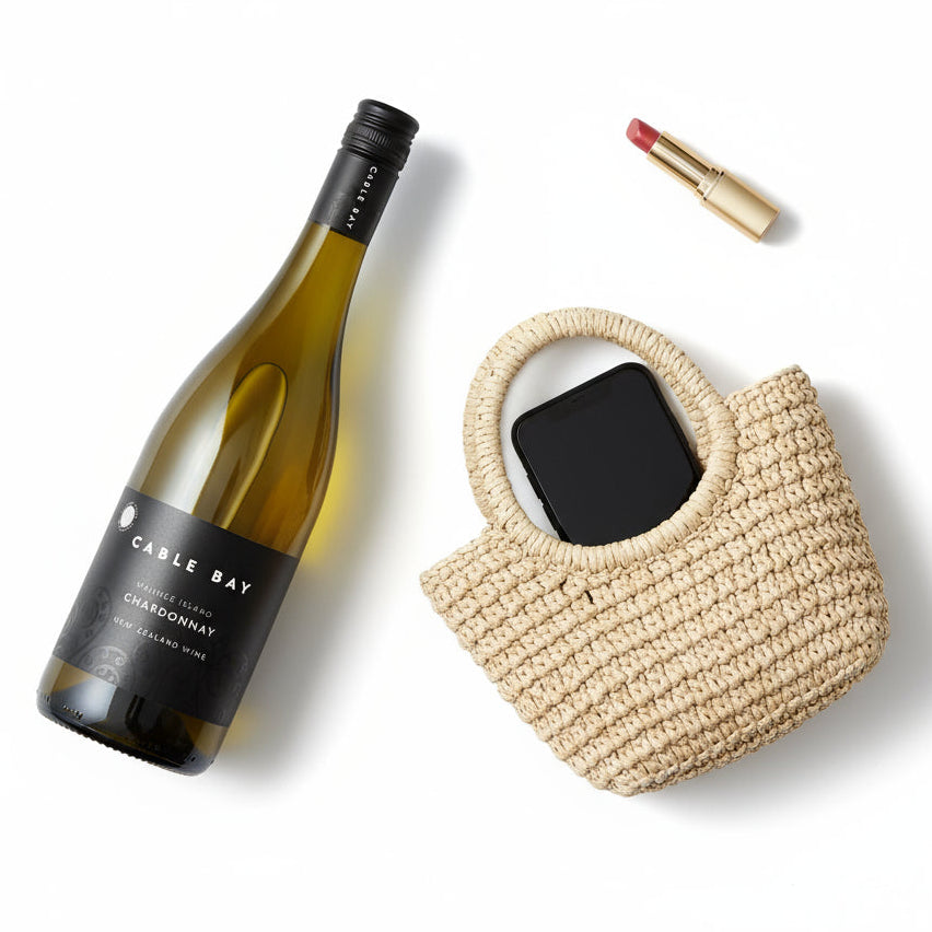 Load image into Gallery viewer, A bottle of Cable Bay Waiheke Island Chardonnay on white background next to a woman's hand bag with mobile and lipstick visible.