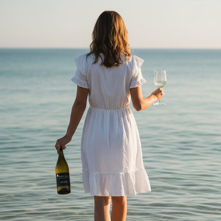 Load image into Gallery viewer, A woman walking into the sea, ankle deep, with a bottle of Cable Bay Waiheke Island Chardonnay wine in her left hand and a wine glass in the right.