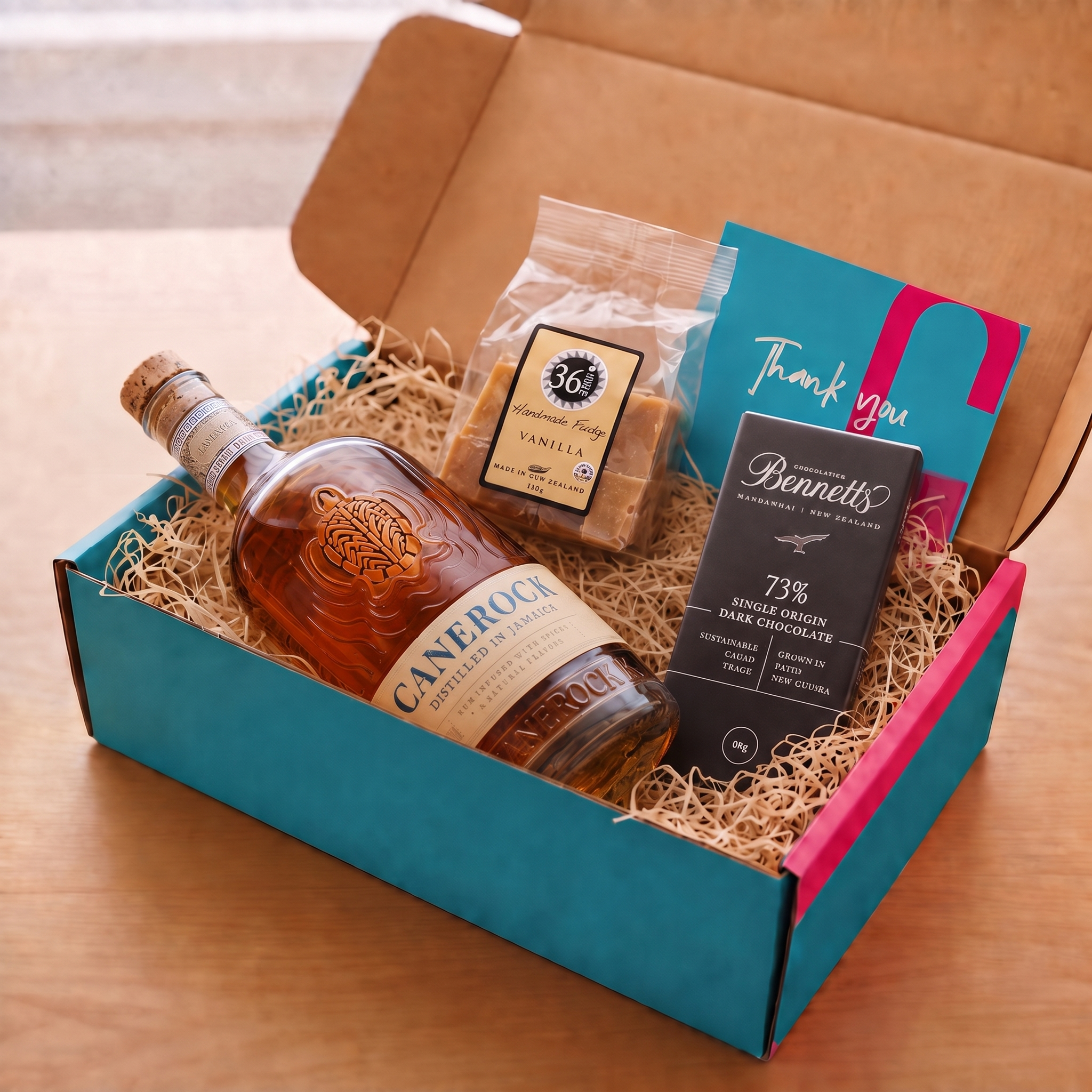 Load image into Gallery viewer, A gift box set on a wooden table containing a bottle of Canerock Jamaican Spiced Rum, a bag of 36 Degrees Vanilla chocolate fudge, a Bennett's 73% single-origin New Zealand dark chocolate bar, and a teal and pink thank you card all in a teal and pink coloured  COVE 27 gift box.