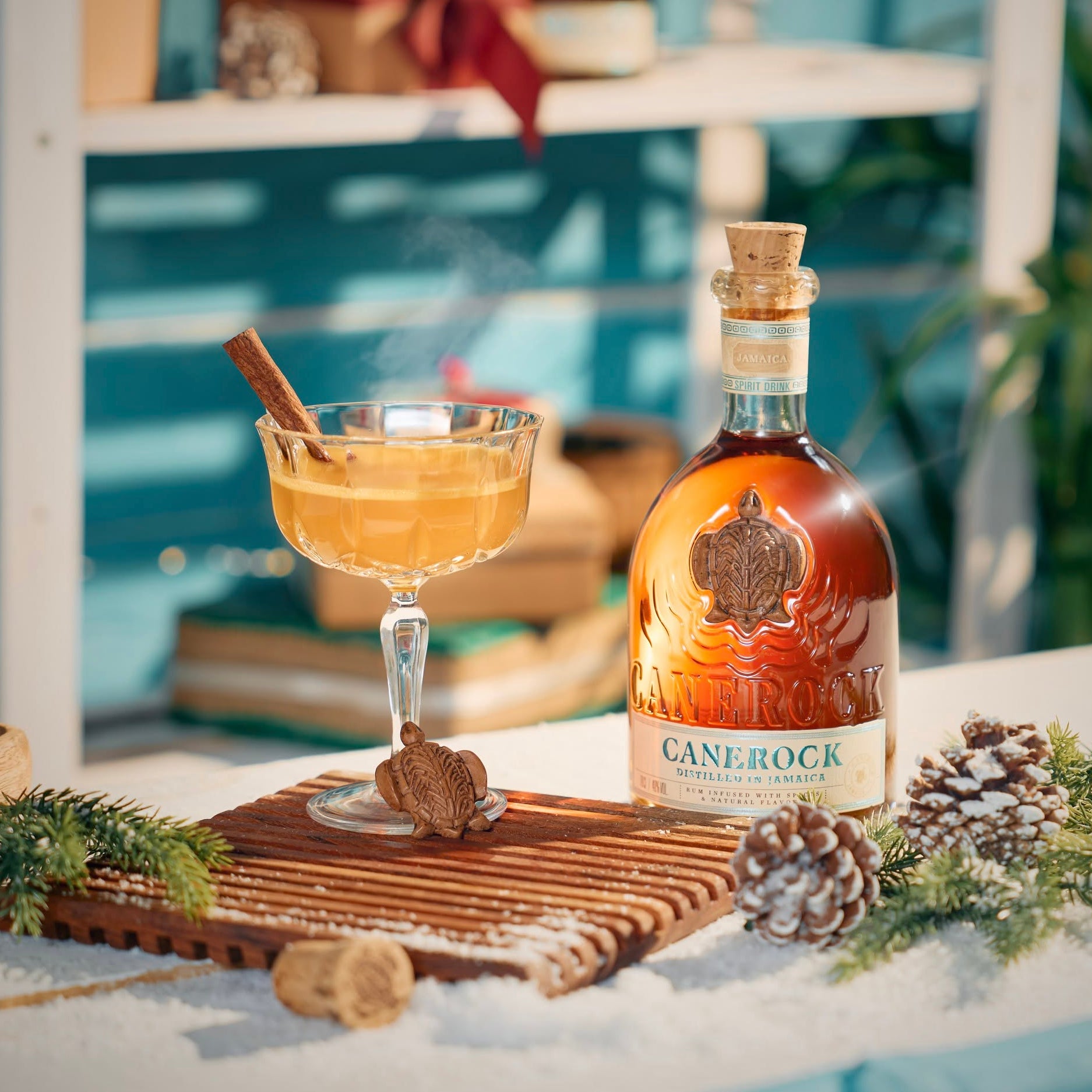 Load image into Gallery viewer, A bottle of Canerock Jamaican Spiced Rum beside a filled coupe glass with a cinnamon stick, styled on a table with pinecones, evergreen branches, and a wooden board in a bright, festive setting.