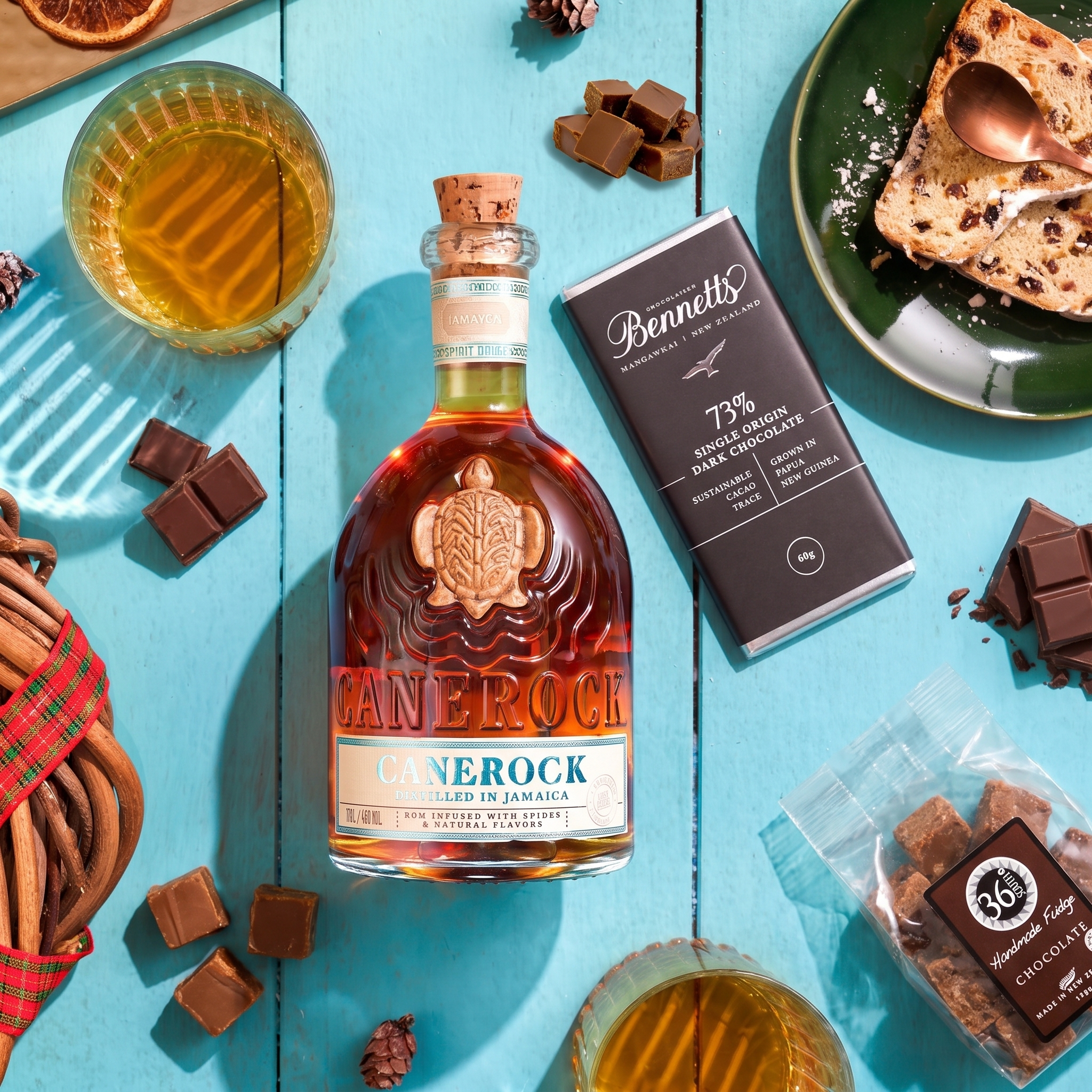 Load image into Gallery viewer, A bottle of Canerock Jamaican Spiced Rum centred on a bright turquoise wooden surface, surrounded by chocolate pieces, a glass of Canerock rum, a Bennett's dark chocolate bar, a packet of 36 Degrees chocolate fudge and a slice of cake. 