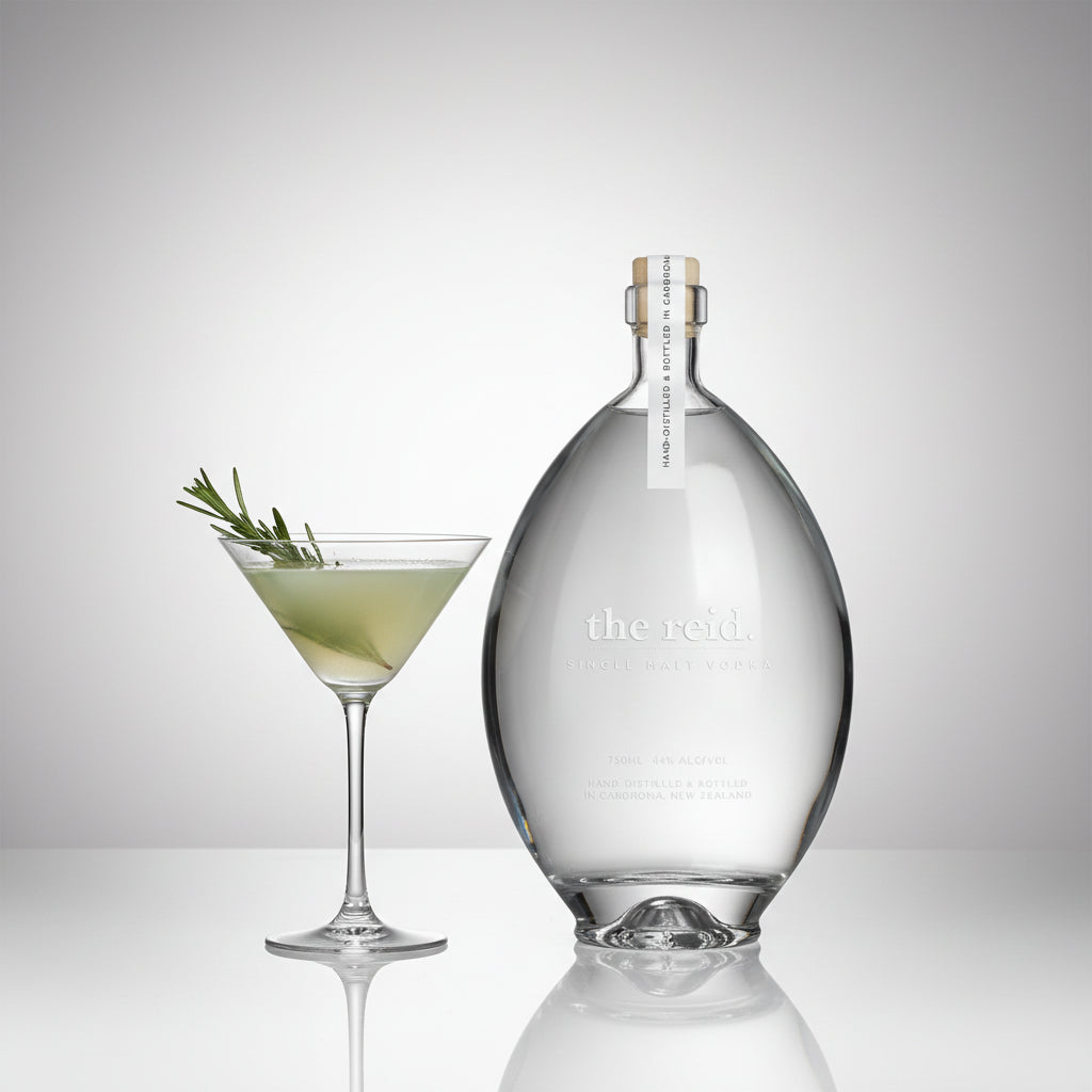 Load image into Gallery viewer, A clear glass bottle labeled 'the reid' single malt vodka stands beside a martini glass filled with pale green cocktail and garnished with a rosemary sprig, on a light grey reflective studio background.