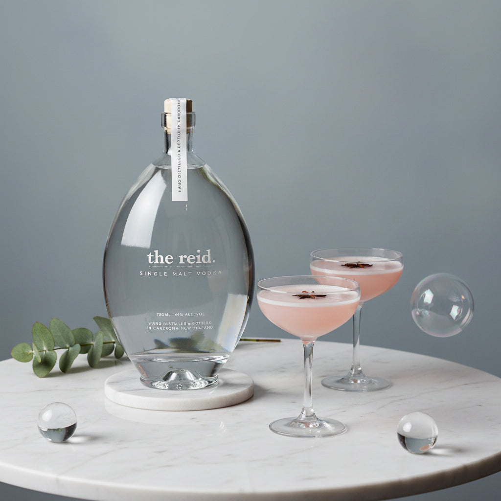 Load image into Gallery viewer, A clear glass decanter of 'the Reid.' single malt vodka on a white marble side table, styled with two pink cocktails in coupe glasses, a green eucalyptus sprig, and decorative glass spheres against a soft gray background.