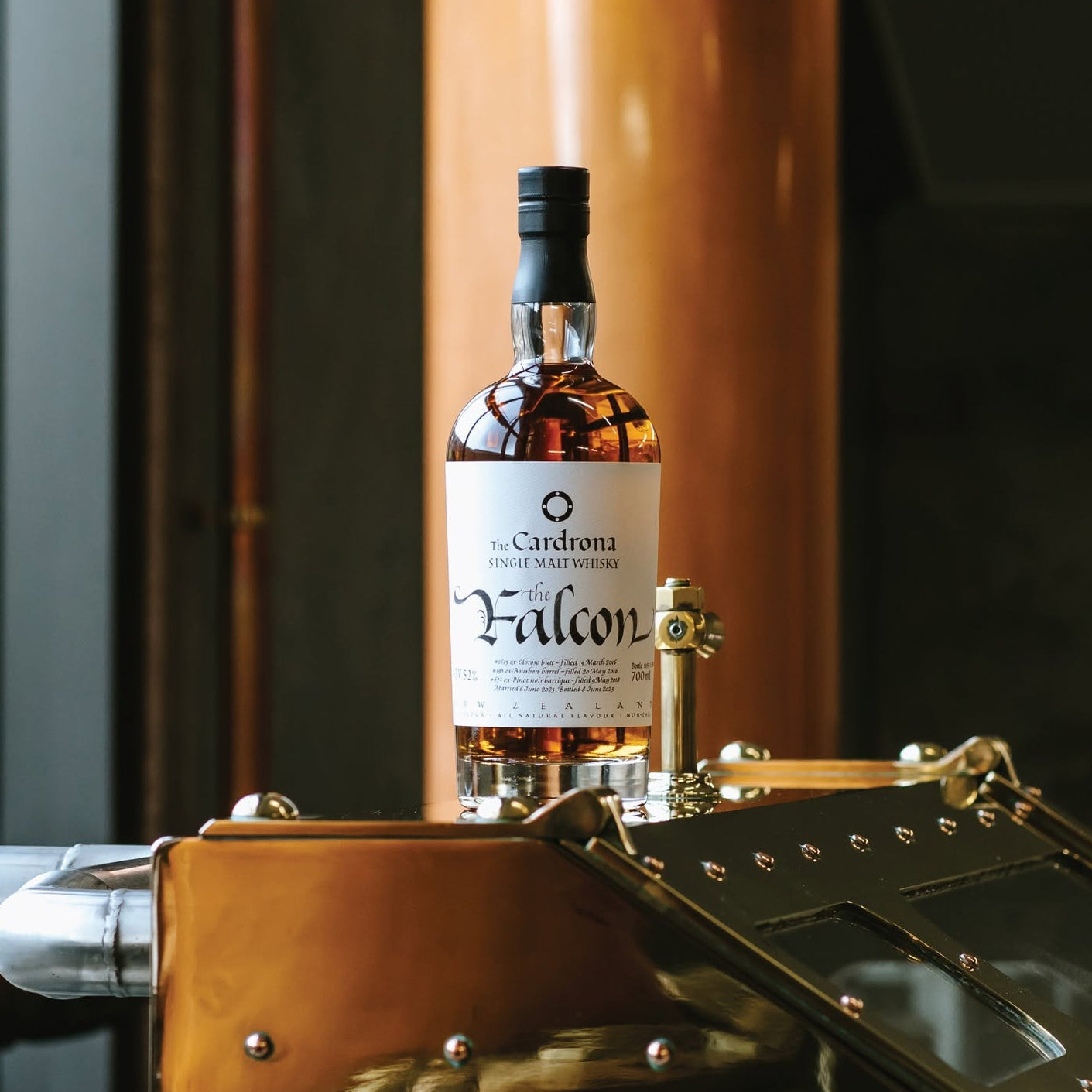 Load image into Gallery viewer, A bottle of Falcon Single Malt Scotch whisky by The Cardona distillery stands on brass distillation equipment inside a whisky distillery, with copper columns and dark metal machinery in the background.