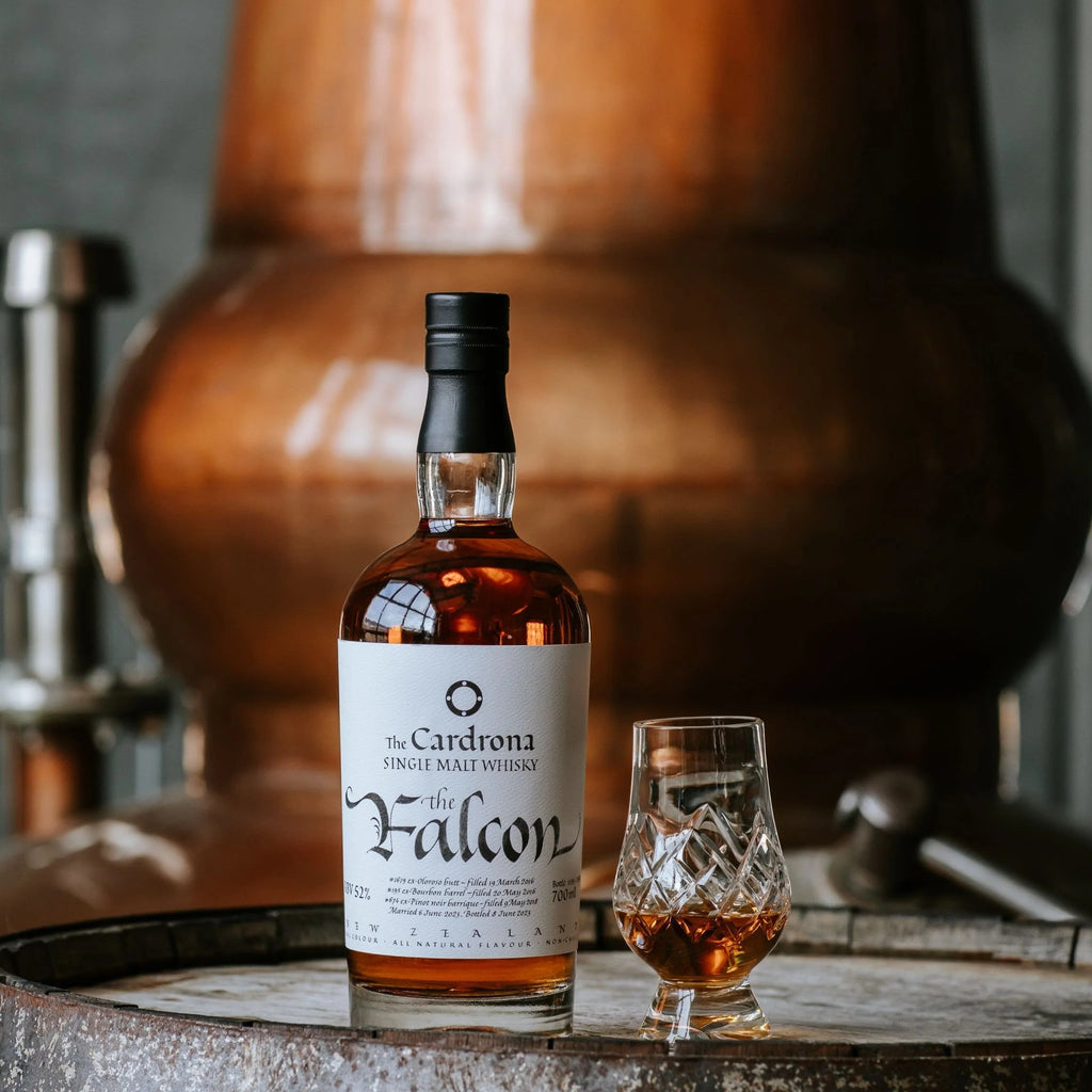 Bottle of The Falcon whisky with a glass on a rustic surface, featuring a large copper still in the background.
